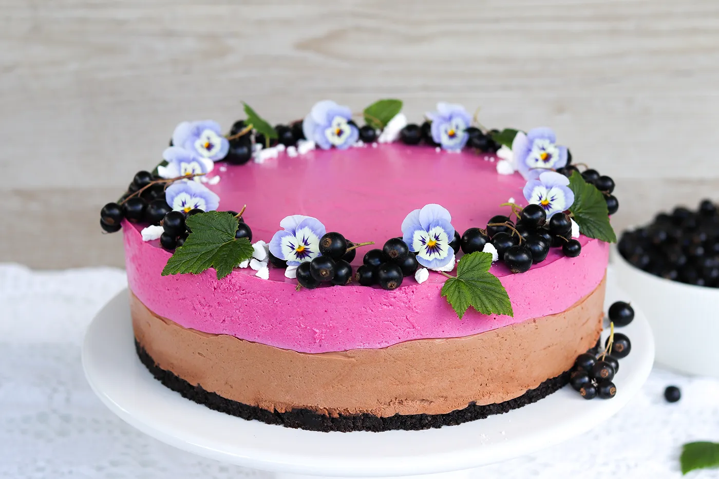 Mustaherukka-suklaajuustokakku, jonka pinnalla on tuoreita marjoja ja orvokkeja.