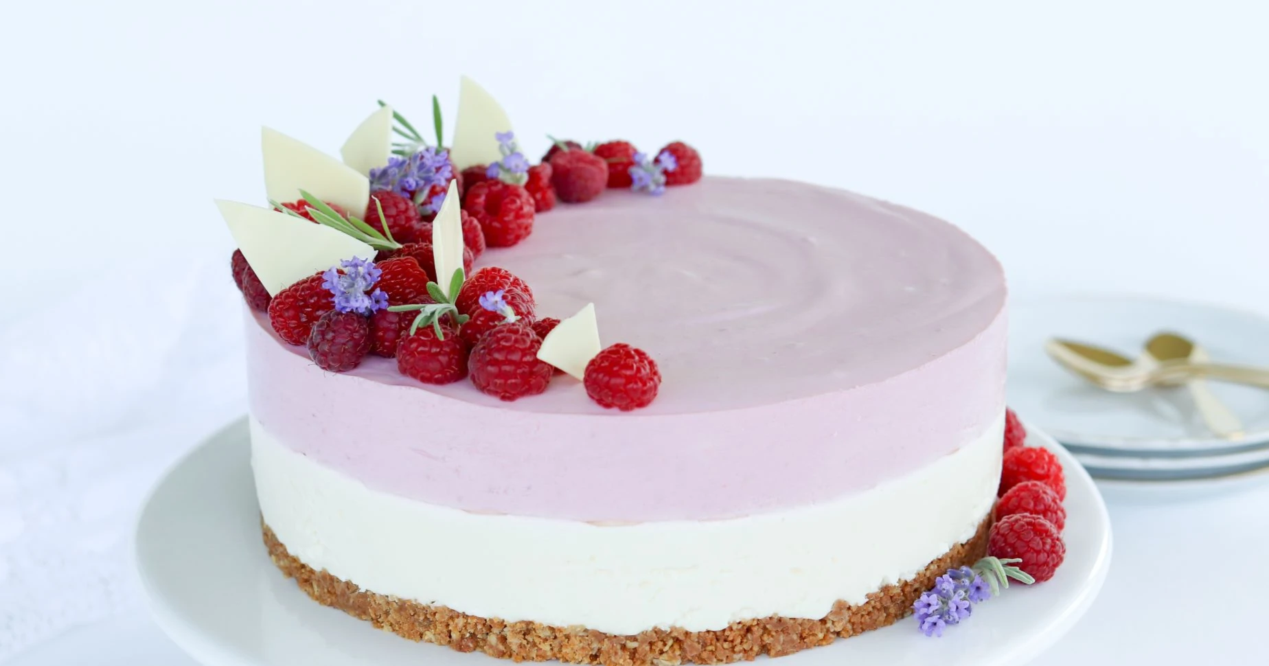 Valkosuklaa-vadelmajuustokakku tarjoilulautasella laventelilla ja vadelmilla koristeltuna.